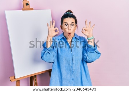 Young hispanic woman standing by painter easel stand looking surprised and shocked doing ok approval symbol with fingers. crazy expression 
