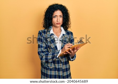 Young latin girl wearing business clothes writing on clipboard skeptic and nervous, frowning upset because of problem. negative person. 