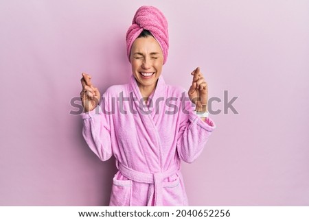 Young blonde woman wearing shower towel cap and bathrobe gesturing finger crossed smiling with hope and eyes closed. luck and superstitious concept. 