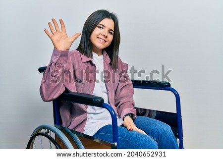 Young brunette woman sitting on wheelchair showing and pointing up with fingers number five while smiling confident and happy. 