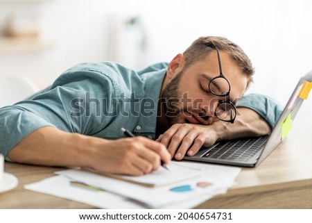 Exhausted millennial man sleeping on his office desk, next to laptop and documents, tired of overworking. Young male workaholic suffering from chronic fatigue at workplace Royalty-Free Stock Photo #2040654731