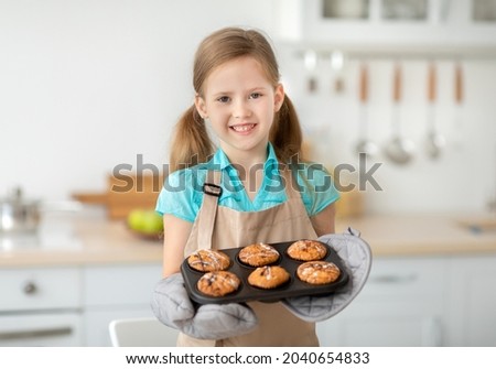 Baking Concept. Portrait of smiling cute little girl holding tray with freshly baked cookies muffins. Positive kid wearing apron, preparing homemade cakes for mother's day. Granny's recipe Royalty-Free Stock Photo #2040654833