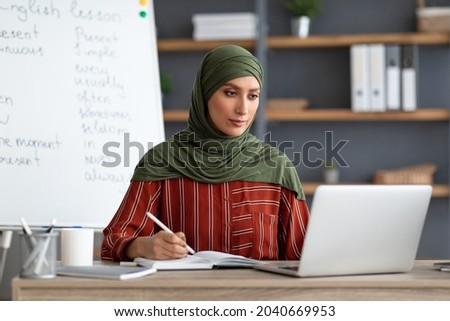 Online Education. Female muslim English teacher sitting at desk using laptop having video conference chat with students and class group. Woman wearing hijab looking at pc screen, writing in notebook