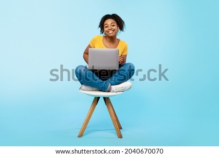 Cheery young black woman working online, sitting on chair and using laptop on blue studio background, full length. Cheerful African American lady surfing internet on portable pc