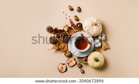 White cup of hot cinnamon tea with an arrangement of dry fall leaves, gourdes, dry apples, nuts, and chestnuts on pastel beige background. Creative fall or Thanksgiving concept. Flat lay.