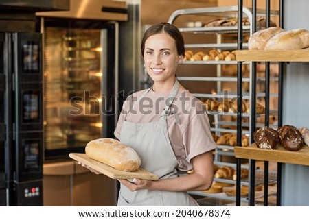 Portrait of happy young female baker in apron standing with fresh loaf of bread by workplace Royalty-Free Stock Photo #2040678374