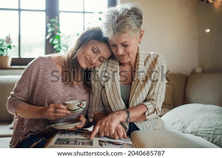 Front view of senior mother with adult daughter indoors at home, looking at family photographs.