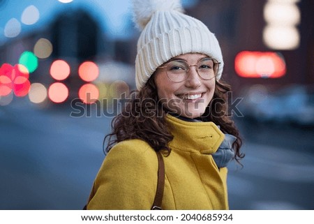 Portrait of happy young woman wearing warm hat with eyeglasses on city street and looking at camera. Beautiful student with cap and winter coat walking at urban road at dusk with copy space.