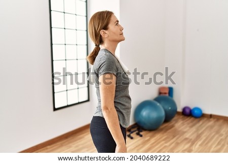Middle age woman wearing sporty look training at the gym room looking to side, relax profile pose with natural face and confident smile. 