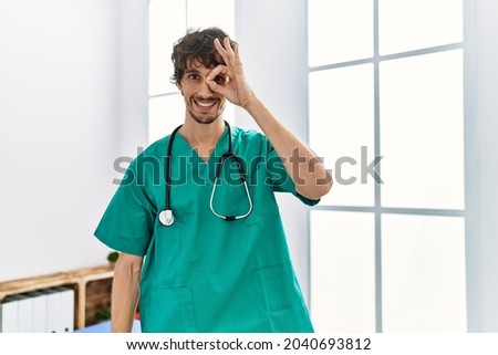 Young hispanic man wearing doctor uniform and stethoscope at clinic smiling happy doing ok sign with hand on eye looking through fingers 