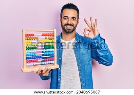 Handsome hispanic man with beard holding traditional abacus doing ok sign with fingers, smiling friendly gesturing excellent symbol 