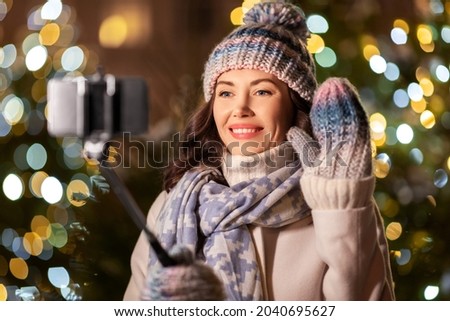 holidays, communication and people concept - portrait of beautiful happy smiling young woman with smartphone and selfie stick taking picture over christmas lights in winter city