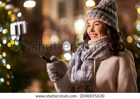 holidays, communication and people concept - portrait of beautiful happy smiling young woman with smartphone and selfie stick taking picture over christmas lights in winter city