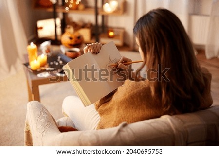 halloween, holidays and leisure concept - young woman with pencil drawing in sketchbook or diary and resting her feet on table at cozy home