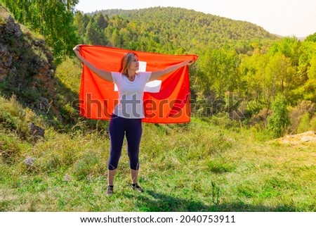 A woman with a European appearance holds the Swiss flag against the backdrop of trees and mountains in sunny weather. Blonde girl with a Swiss flag in her hands