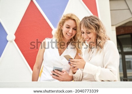 Two pretty young woman watching video on phone. Happy female person outdoors. Summer city business portrait. Lady calling friends. Blonde hair. Cute adult student at town