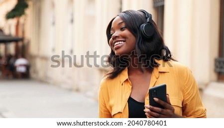 Charming multiracial lady wearing headphones standing in urban city streets and listening music. Young female teenager dancing and shaking head while enjoying of the music. Entertainment concept 