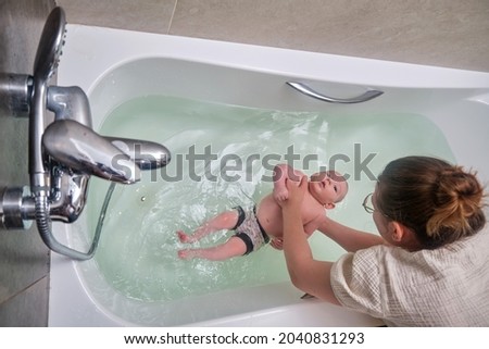 Mom and son are engaged in infant swimming in the home bathroom. A woman helps a newborn baby to swim in a large white bath.