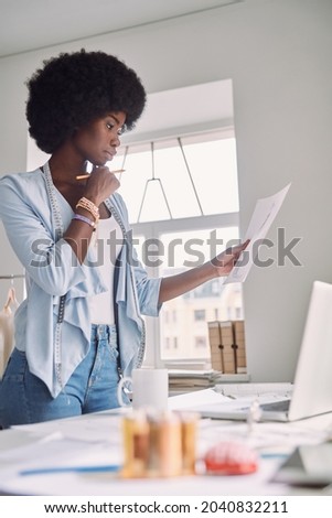 Beautiful young African woman analyzing fashion design sketch while standing near her desk in office