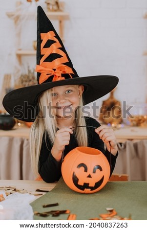 Portrait of a little blonde girl with long hair in a witch costume with huge witch hat, who is holding a pumpkin-shaped candy basket in her hands. Halloween concept. Space for text. High quality photo