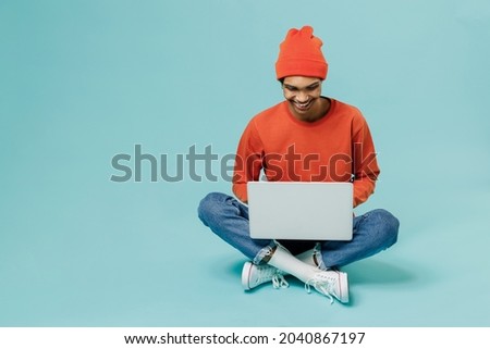Full body young smiling satisfied happy african american man 20s in orange shirt hat sitting on floor hold use work on laptop pc computer isolated on plain pastel light blue background studio portrait