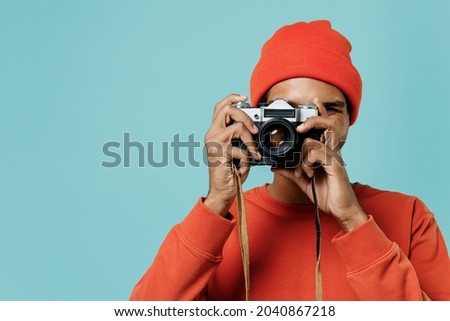 Young smiling happy african american man 20s in orange shirt hat taking photo picture on retro vintage photo camera isolated on plain pastel light blue background studio. People lifestyle concept.