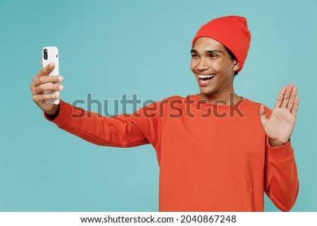 Young happy african american man in orange shirt hat doing selfie shot on mobile cell phone post photo on social network waving hand say hello isolated on plain pastel light blue background studio.