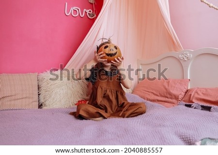 Cute little girl playing with Halloween pumpkin on bed in her pink room. Girl looking at camera and smiling. halloween party at home. selective focus.