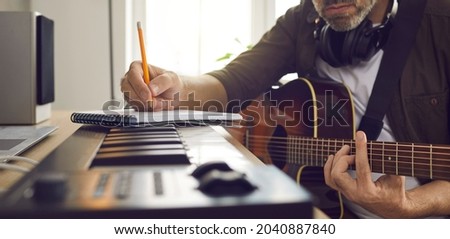 Musician writing music. Songwriter who's following his passion sitting by electronic keyboard in home studio, holding pencil, picking guitar strings, thinking of lyrics for new song. Banner background Royalty-Free Stock Photo #2040887840