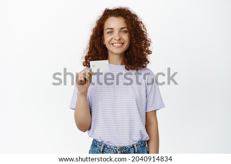 Stylish smiling redhead woman, showing her credit card and looking happy, become bank client, standing in casual t-shirt over white background