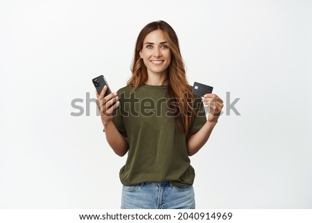 Smiling brunette woman showing her credit discount card, holding smartphone mobile phone, looking pleased at camera, standing against white background