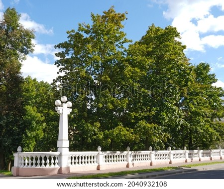 The fence of the old park, lanterns, green trees. Clouds are floating across the blue sky.
