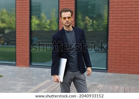 Handsome man in suit is standing near his office. He is holding a laptop and looking at the camera. 
Copy space