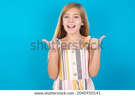 Optimistic beautiful caucasian little girl wearing striped dress over blue background showing thumbs up with positive emotions. Quality and recommendation concept.