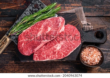 Uncooked Raw Shoulder Top Blade or flat iron beef meat steaks on a wooden butcher board with meat cleaver. Dark wooden background. Top View Royalty-Free Stock Photo #2040963074