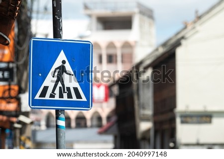 A pedestrian crosswalk signpost symbol in blue and triangle logo on the street with city as blurred background. Transportation sign object photo.