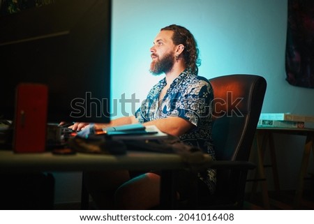 Focused bearded man use computer against teal light sitting on chair