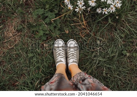 women's legs in a long dress and green sneakers on the autumn withered grass and a daisy bush. autumn mood picture. selective focus