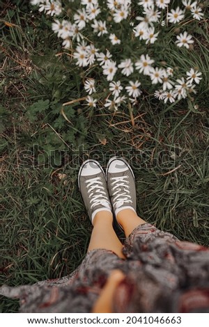 women's legs in a long dress and green sneakers on the autumn withered grass and a daisy bush. autumn mood picture. selective focus
