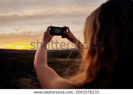 Rear view of young woman holding smartphone taking picture of morning sunrise against horizon
