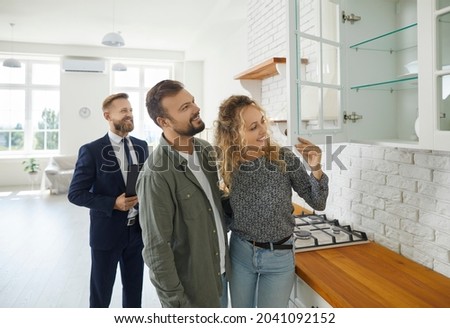 Boyfriend and girlfriend buying house. First time buyers or future tenants meeting real estate agent and looking around new home. Husband and wife looking at good quality modern wooden kitchen cabinet Royalty-Free Stock Photo #2041092152