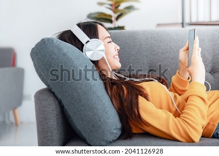 Happy asian woman using mobile smartphone and headphones while listening online music media entertainment relaxation in a sofa at home. Royalty-Free Stock Photo #2041126928