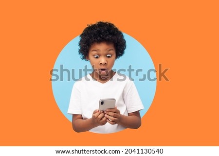 shocked boy with curly hair reading message on smartphone and shocked, surprised by mobile application, using cellphone. indoor studio shot isolated in a round hole on orange background
