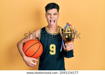 Young hispanic man wearing basketball uniform holding ball and prize sticking tongue out happy with funny expression. 