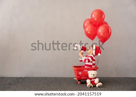 Happy child wearing Christmas costume. Kid sitting in shopping cart with red balloons. Funny child playing. Kid speaking loudspeaker. Black friday sale concept