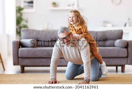 Cute little girl granddaughter playing and having fun with active positive grandfather, senior man grandpa giving piggyback to excited child and smiling at camera while taking care of kid at home Royalty-Free Stock Photo #2041162961