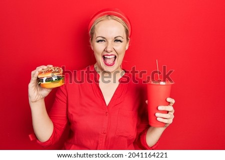 Young blonde woman eating a tasty classic burger with fries and soda smiling and laughing hard out loud because funny crazy joke. 