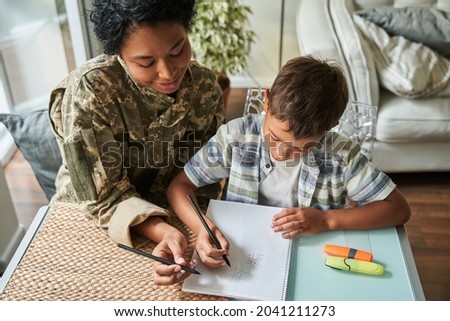 Woman wearing military outfit sitting at the table with her little son and drawing