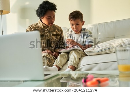 Woman wearing military jacket talking with her little son while sitting at the sofa
