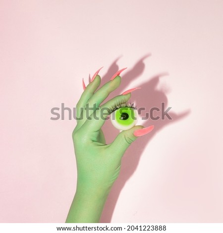 Creative layout with green painted hand with bright pink nails holding green eyeball against pastel pink background. Halloween celebration idea. Minimal flat lay concept. Royalty-Free Stock Photo #2041223888